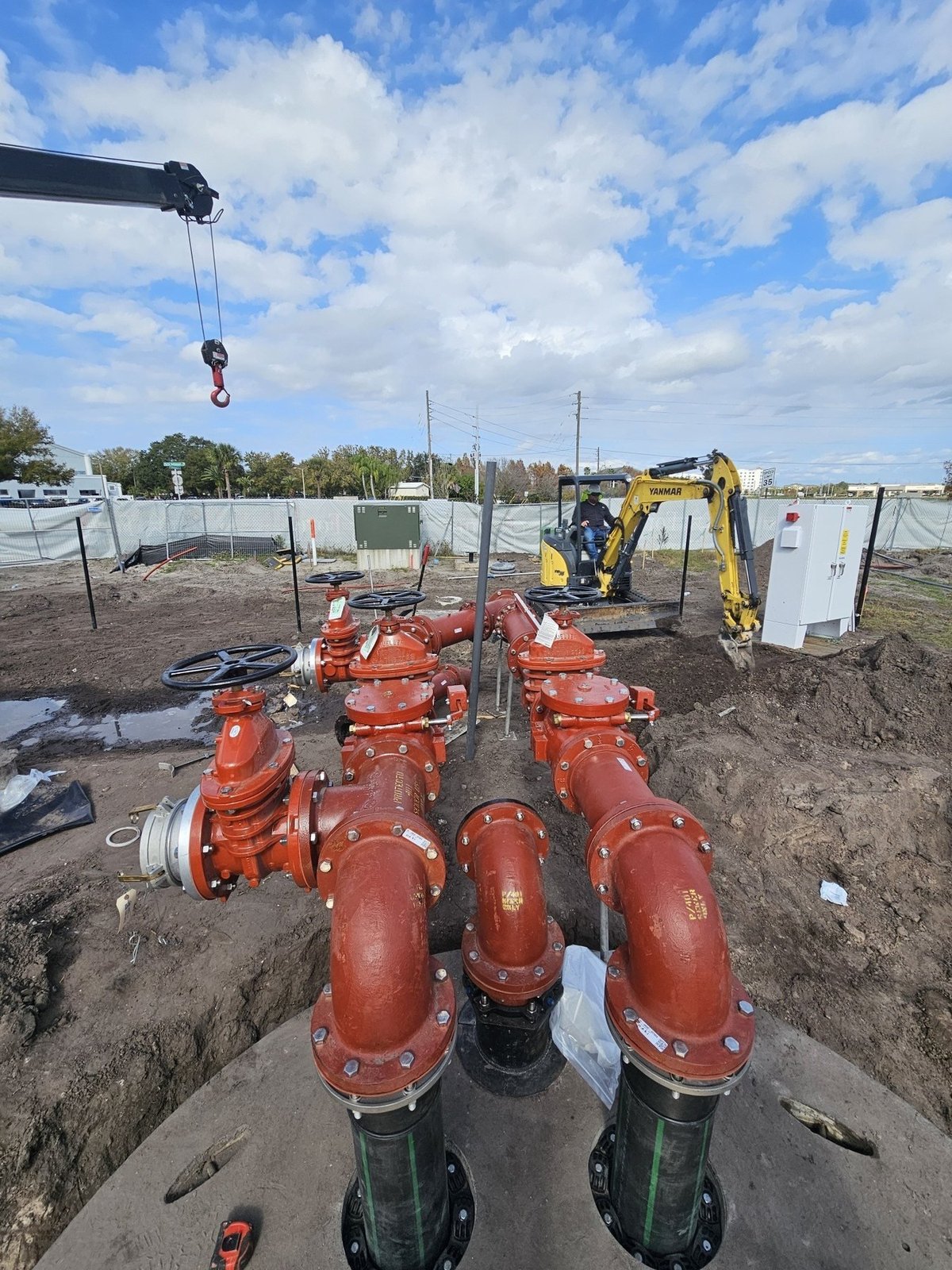 Industrial construction site showing large red pipe infrastructure with yellow excavator and crane equipment under clear sky